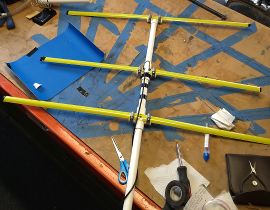 An overhead shot of a yagi antenna, made with a PVC pipe and metal tape measure, lying on a work table.